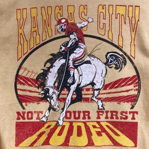 May include: A light yellow t-shirt featuring a graphic of a football player riding a bucking bronco. The words "Kansas City" are arched above the image, with "Not Our First Rodeo" and "Rodeo" below. The design is in red and yellow.