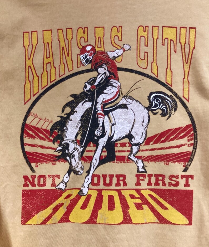May include: A light yellow t-shirt featuring a graphic of a football player riding a bucking bronco. The words "Kansas City" are arched above the image, with "Not Our First Rodeo" and "Rodeo" below. The design is in red and yellow.
