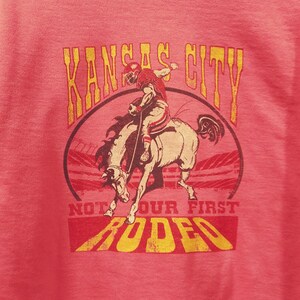 May include: A red t-shirt with a distressed graphic featuring a football player riding a bucking bronco. The text "Kansas City" arches above the image, with "Not Our First Rodeo" below.