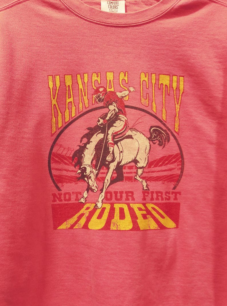 May include: A red t-shirt with a distressed graphic featuring a football player riding a bucking bronco. The text "Kansas City" arches above the image, with "Not Our First Rodeo" below.