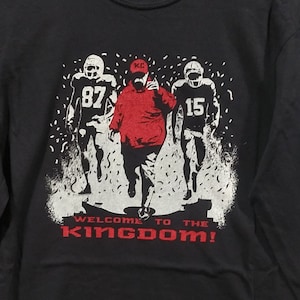 May include: Black sweatshirt with a graphic of three football players in white and red. The center figure is in red, wearing a KC hat. The text "WELCOME TO THE KINGDOM!" is in red at the bottom.