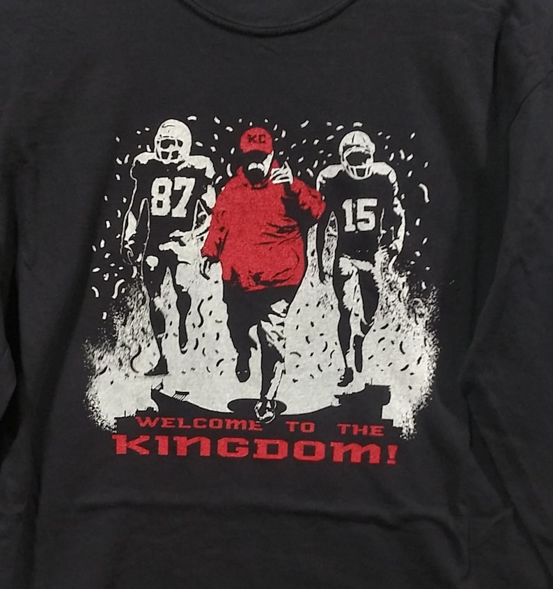 May include: Black sweatshirt with a graphic of three football players in white and red. The center figure is in red, wearing a KC hat. The text "WELCOME TO THE KINGDOM!" is in red at the bottom.