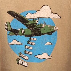 May include: A tan t-shirt featuring a cartoon illustration of a green airplane dropping bombs. The plane is set against a blue circle with white clouds. The bombs have the letter "F" on them.