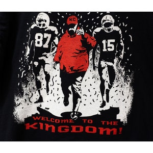 May include: Black t-shirt with a graphic of three football figures in red and white. The central figure wears a red cap with "KC" on it. The text "WELCOME TO THE KINGDOM!" is printed in red at the bottom.
