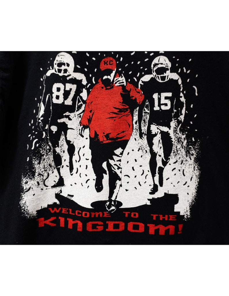 May include: Black t-shirt with a graphic of three football figures in red and white. The central figure wears a red cap with "KC" on it. The text "WELCOME TO THE KINGDOM!" is printed in red at the bottom.