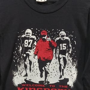 May include: Black crew neck t-shirt with a graphic of three football players in white and black uniforms. The central figure wears a red hoodie. The text "WELCOME TO THE KINGDOM!" is printed in red.