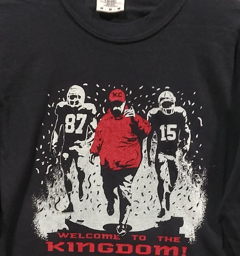 May include: Black crew neck t-shirt with a graphic of three football players in white and black uniforms. The central figure wears a red hoodie. The text "WELCOME TO THE KINGDOM!" is printed in red.