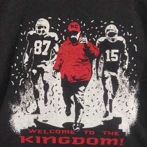 May include: Black sweatshirt with a graphic of three football figures in white and red. The center figure is in red, with the text "WELCOME TO THE KINGDOM!" in red at the bottom. The figures are set against a white background with confetti.