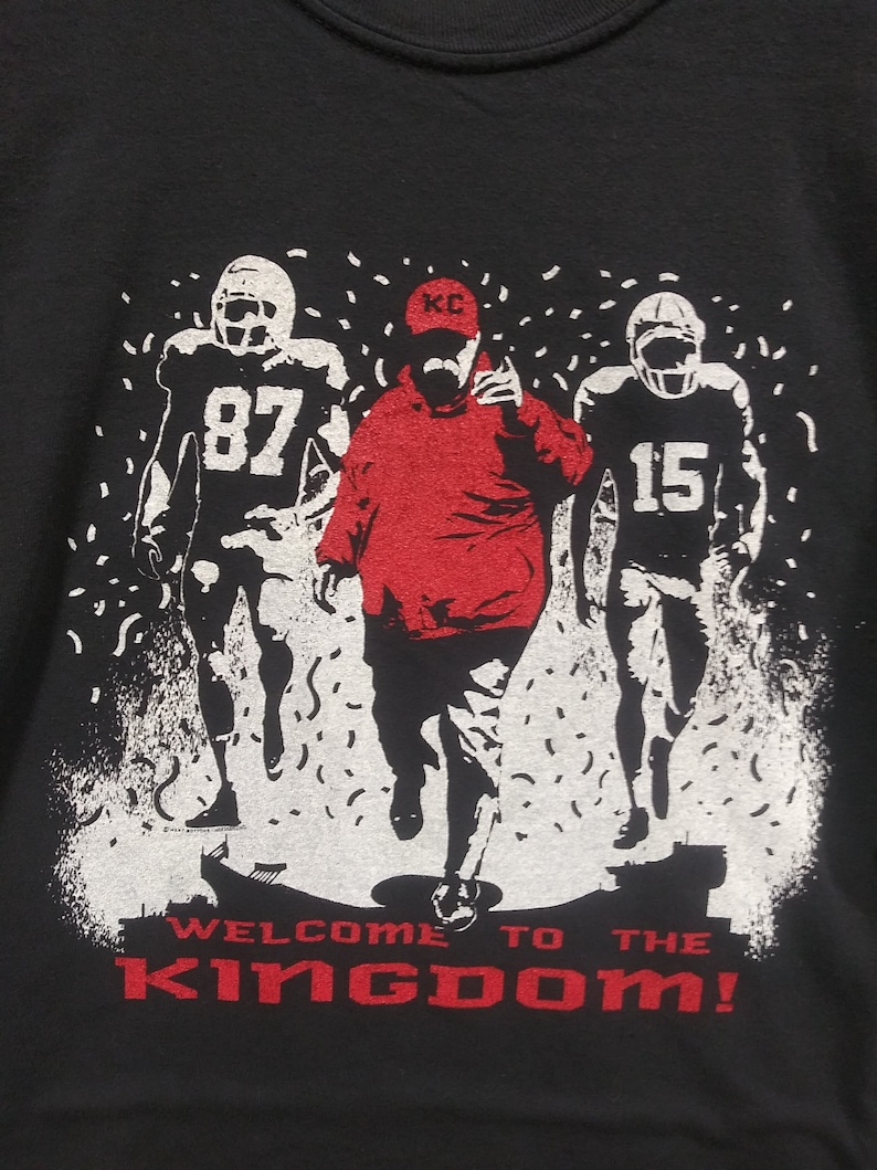 May include: Black sweatshirt with a graphic of three football figures in white and red. The center figure is in red, with the text "WELCOME TO THE KINGDOM!" in red at the bottom. The figures are set against a white background with confetti.