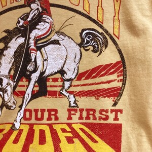 May include: Mustard yellow t-shirt featuring a graphic of a football player riding a horse. The design includes the words "Kansas City" and "Our First Rodeo" in red and yellow lettering. The shirt has a vintage, distressed look.