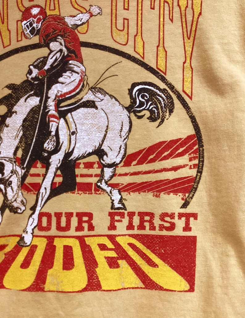 May include: Mustard yellow t-shirt featuring a graphic of a football player riding a horse. The design includes the words "Kansas City" and "Our First Rodeo" in red and yellow lettering. The shirt has a vintage, distressed look.