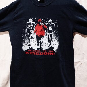 May include: Black t-shirt featuring a graphic of three football players, one in a red jacket and hat, with the text "WELCOME TO THE KINGDOM!" in red. The players are in white, with the numbers 87 and 15 visible.