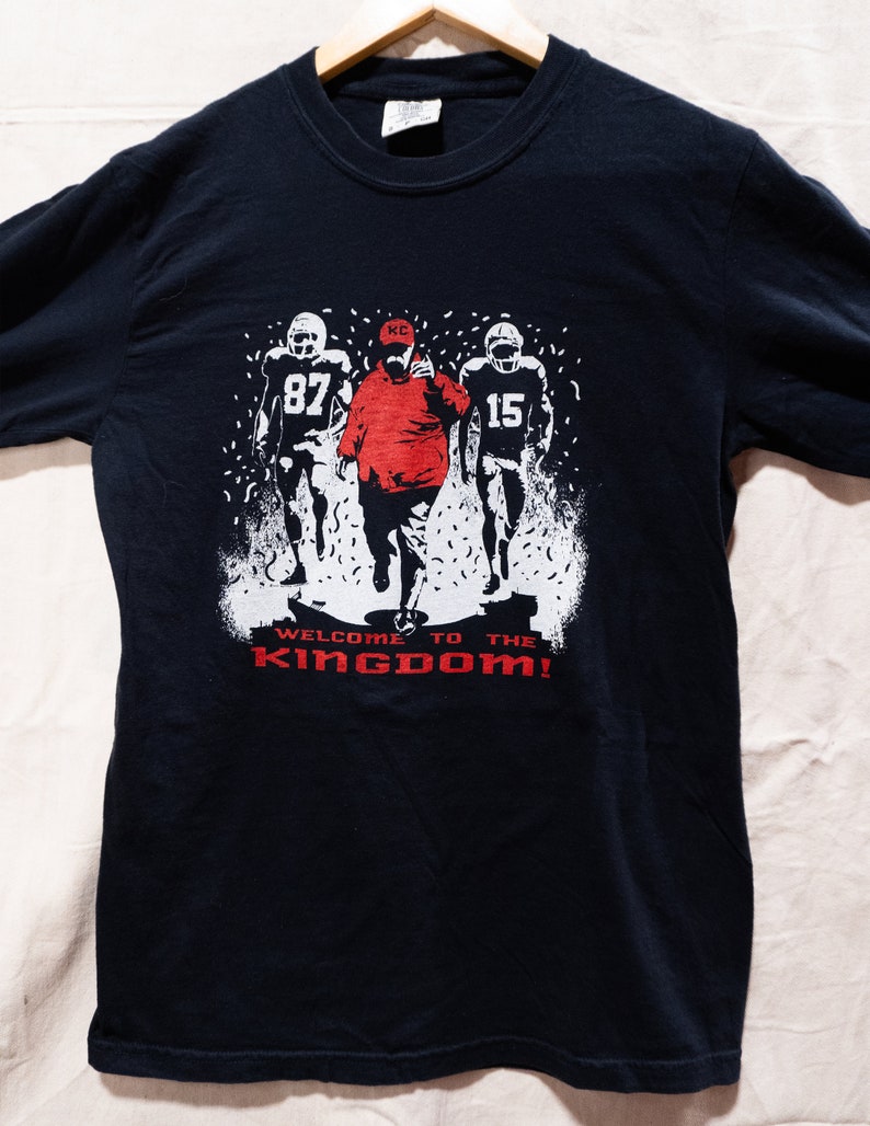 May include: Black t-shirt featuring a graphic of three football players, one in a red jacket and hat, with the text "WELCOME TO THE KINGDOM!" in red. The players are in white, with the numbers 87 and 15 visible.