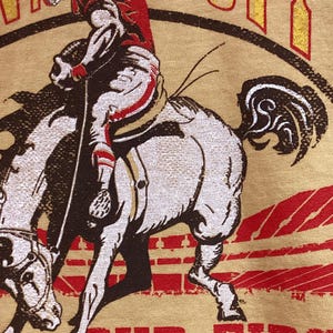 May include: A tan t-shirt featuring a vintage-style graphic of a football player riding a horse. The player is wearing a red and white uniform and helmet. The text "OUR FIRST" is printed in red at the bottom of the image.