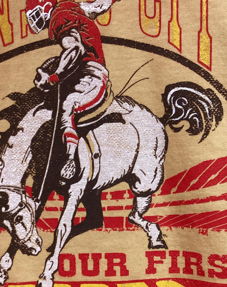 May include: A tan t-shirt featuring a vintage-style graphic of a football player riding a horse. The player is wearing a red and white uniform and helmet. The text "OUR FIRST" is printed in red at the bottom of the image.