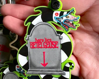 May include: A green, black, and white Beetlejuice keychain. The keychain features a tombstone design with the words "here lies BETELGEUSE" and a red arrow. The keychain also has a cartoon-style monster head with a red and blue color scheme.