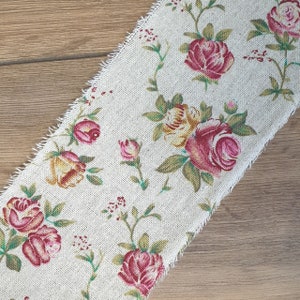 May include: A beige fabric with a floral pattern featuring pink, yellow, and green roses. The fabric has a frayed edge.