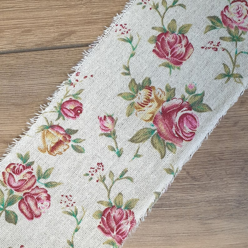 May include: A beige fabric with a floral pattern featuring pink, yellow, and green roses. The fabric has a frayed edge.