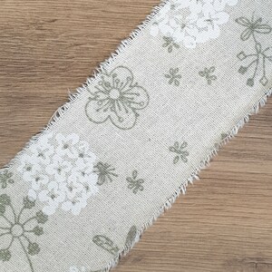 May include: A beige fabric ribbon with a white and green floral pattern. The ribbon has a frayed edge.