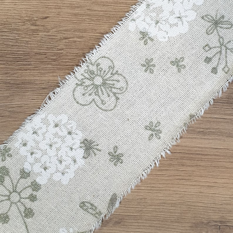 May include: A beige fabric ribbon with a white and green floral pattern. The ribbon has a frayed edge.