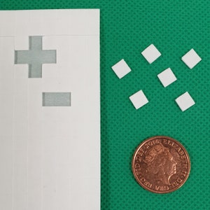 May include: A white sheet of paper with a plus sign and a minus sign cut out of it. There are several small white squares on a green surface.