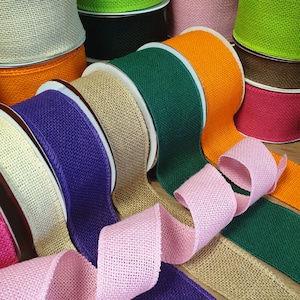 May include: A variety of burlap ribbon rolls in different colors, including natural, pink, purple, green, orange, and red. The rolls are arranged in a semi-circle, with some of the ribbon unrolled and overlapping.