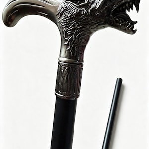 May include: A walking stick featuring a metallic silver-toned handle sculpted into the shape of a snarling wolf head with bared teeth. The handle connects to a smooth, matte black shaft via a decorative, textured silver-toned collar band.