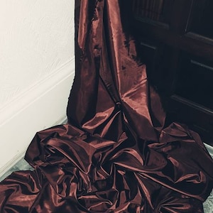 May include: A deep burgundy satin fabric draped over a wooden floor. The fabric is gathered and folded, creating soft, flowing lines.