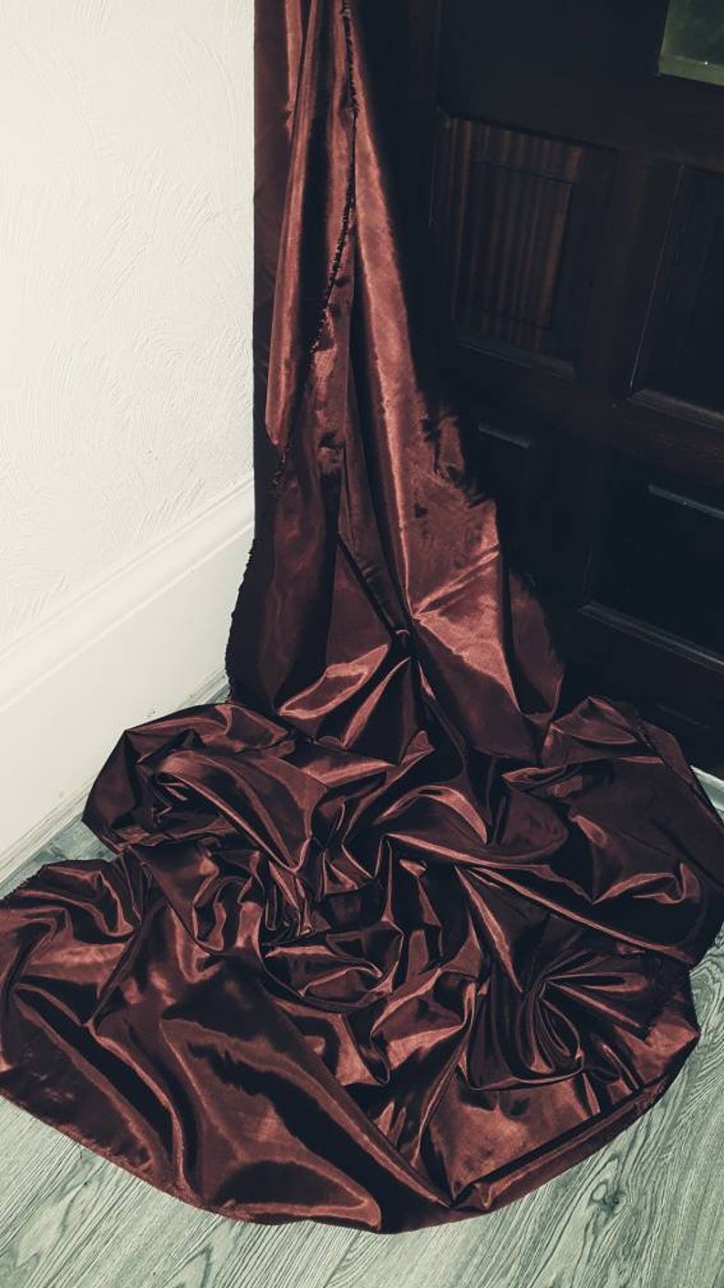May include: A deep burgundy satin fabric draped over a wooden floor. The fabric is gathered and folded, creating soft, flowing lines.