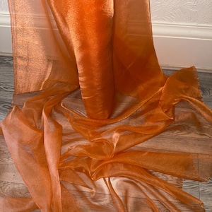 May include: A length of orange sheer fabric, with a shimmering quality, is draped across a wooden surface. The translucent material allows light to pass through, creating a play of light and shadow. The fabric's folds and waves add texture and movement. The colour is a vibrant orange.