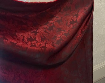 May include: Red paisley patterned fabric with a satin-like sheen. The fabric is folded and draped over a white surface.