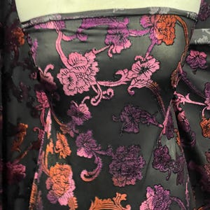 Op de afbeelding: Zwarte, doorschijnende stof met een bloemenpatroon in tinten roze, paars en oranje. De stof heeft een subtiele glitterafwerking.