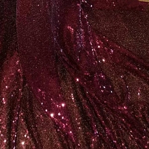 May include: Close-up of a burgundy fabric with sequins. The sequins are small and densely packed, creating a shimmering effect.