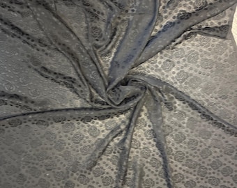 May include: Black fabric with a repeating pattern of roses and dots. The fabric is draped and folded, creating shadows and texture. The material appears to be soft and flowing, suitable for clothing or decorative projects.