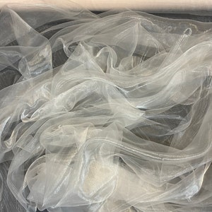 Könnte beinhalten: Eine Rolle weißen, durchsichtigen Stoffes mit schimmerndem Finish. Der Stoff ist gefaltet und auf einer dunklen Oberfläche drapiert.