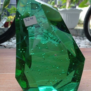 Op de afbeelding: Een groot, groen, gefacetteerd glazen beeldhouwwerk met ingesloten luchtbellen. Het beeldhouwwerk is gevormd als een grillige, puntige rots. Een label op het beeldhouwwerk luidt "3/2 kg".