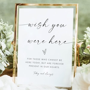 May include: A gold-framed sign with the words "wish you were here" in elegant script. Below, the text reads, "FOR THOSE WHO CANNOT BE HERE TODAY, BUT ARE FOREVER PRESENT IN OUR HEARTS." The sign is surrounded by greenery and white fabric.