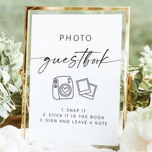 Puede incluir: Un letrero con marco dorado con las palabras "PHOTO guestbook" en escritura elegante. Debajo, ilustraciones de cámara y fotos acompañan las instrucciones: "1. SNAP IT, 2. STICK IT IN THE BOOK, 3. SIGN AND LEAVE A NOTE." El letrero está sobre una superficie de madera con vegetación.
