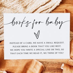 May include: A white card with the text "books for baby" in elegant script, with a heart symbol below. The card's message requests guests to bring a book and write a special line. The card rests on a wooden surface with tan fabric.
