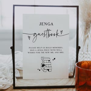 Puede incluir: Un letrero enmarcado con el texto "JENGA guestbook" en escritura negra. El letrero anima a los invitados a firmar una pieza de Jenga con buenos deseos. Una pila de bloques de Jenga está ilustrada en la parte inferior. Un vaso decorativo y un jarrón están en el fondo.