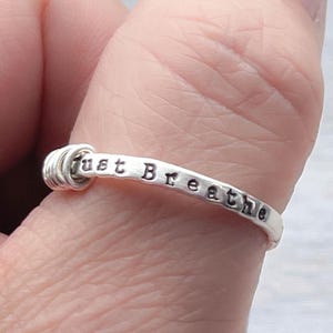May include: A silver ring with the words "just breathe" stamped on the band. The ring has a simple, elegant design, with a coiled detail at the top. The ring is shown on a person's finger.