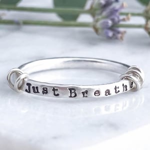 A sterling silver fidget ring with the words 'Just Breathe' hand-stamped on the front, resting on a white marble surface with florals behind