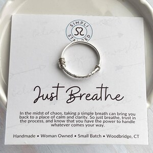 Pu&ograve; includere: Un anello d'argento con una texture martellata e dettagli avvolti, su una carta bianca. La carta presenta la scritta "Just Breathe" in corsivo, insieme al testo "Handmade Woman Owned Small Batch Woodbridge, CT".