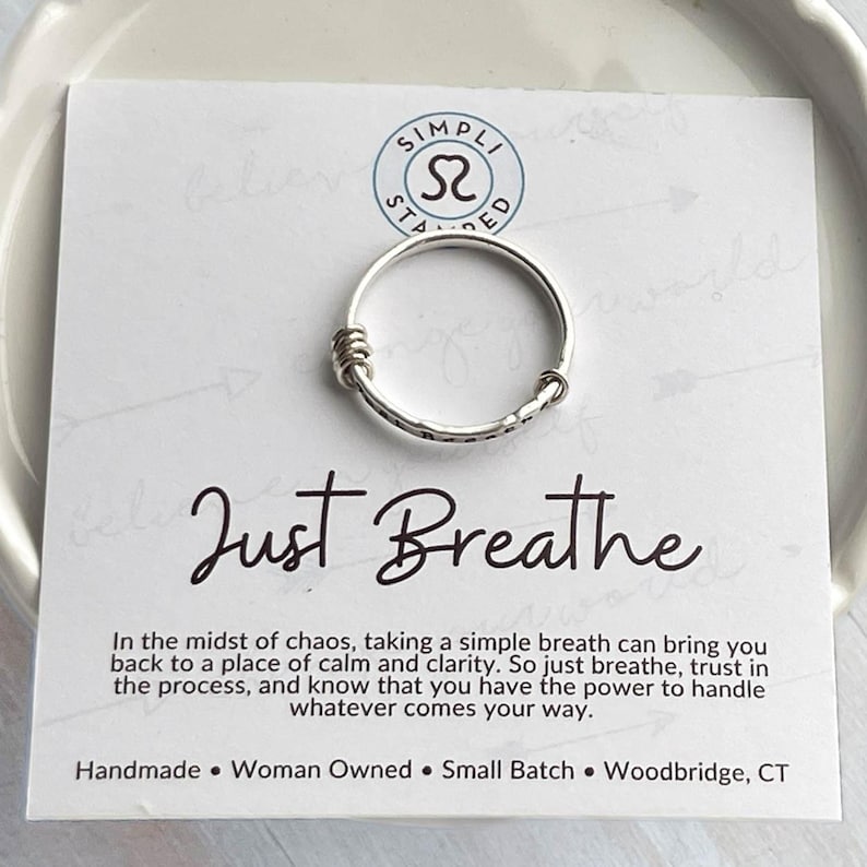 Pu&ograve; includere: Un anello d'argento con una texture martellata e dettagli avvolti, su una carta bianca. La carta presenta la scritta "Just Breathe" in corsivo, insieme al testo "Handmade Woman Owned Small Batch Woodbridge, CT".