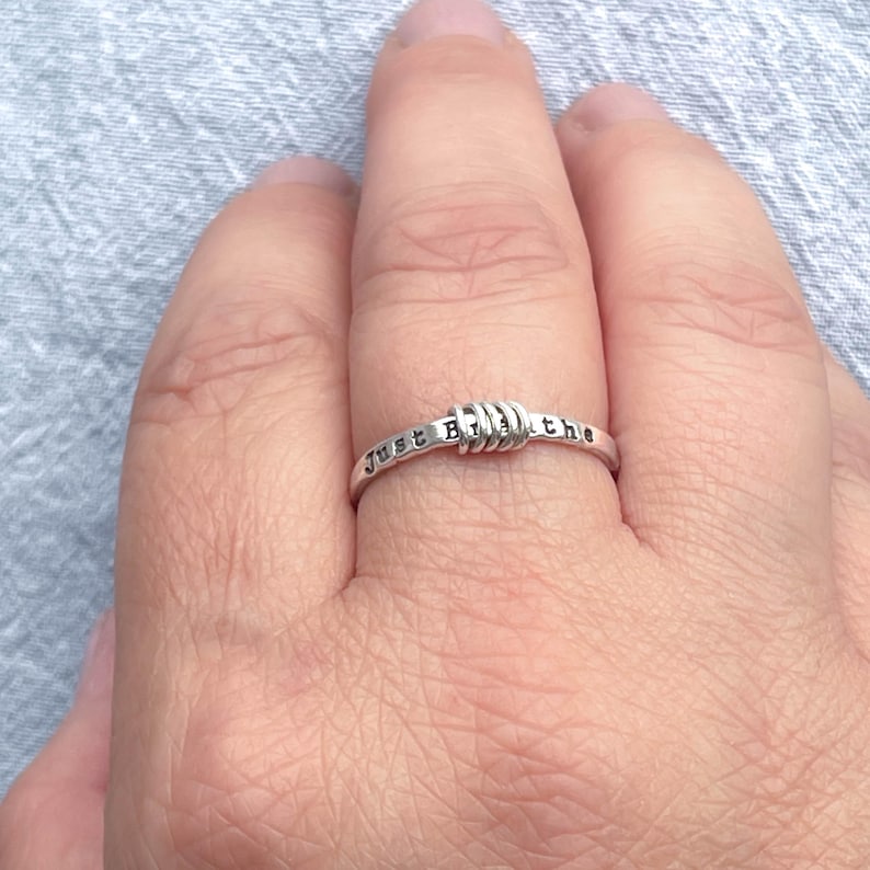 Pu&ograve; includere: Un anello d'argento con la scritta "Just Breathe". L'anello presenta un design unico con un centro avvolto. L'anello &egrave; mostrato sul dito di una persona.