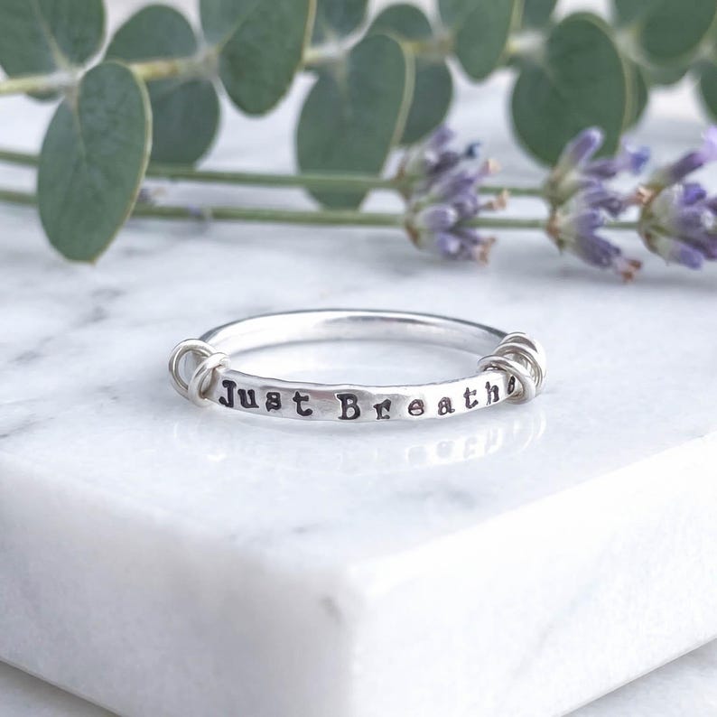 A sterling silver fidget ring with the words 'Just Breathe' hand-stamped on the front, resting on a white marble surface with florals behind