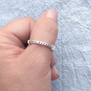 Pu&ograve; includere: Un anello d'argento con la scritta "Just Breathe". L'anello &egrave; indossato su un dito e il testo &egrave; chiaramente visibile. L'anello ha un design semplice ed elegante. L'anello &egrave; d'argento.