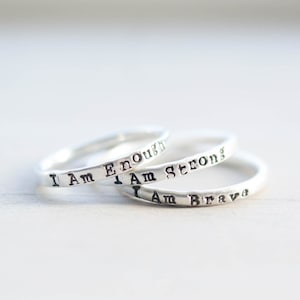 May include: Three silver rings stacked on top of each other. The rings are inscribed with the words "I Am Enough", "I Am Strong", and "I Am Brave".
