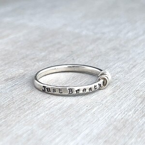 Pu&ograve; includere: Un anello d'argento con le parole "Just Breathe" incise. L'anello presenta un design minimalista con un elemento decorativo su un lato. L'anello &egrave; mostrato su una superficie grigio chiaro.