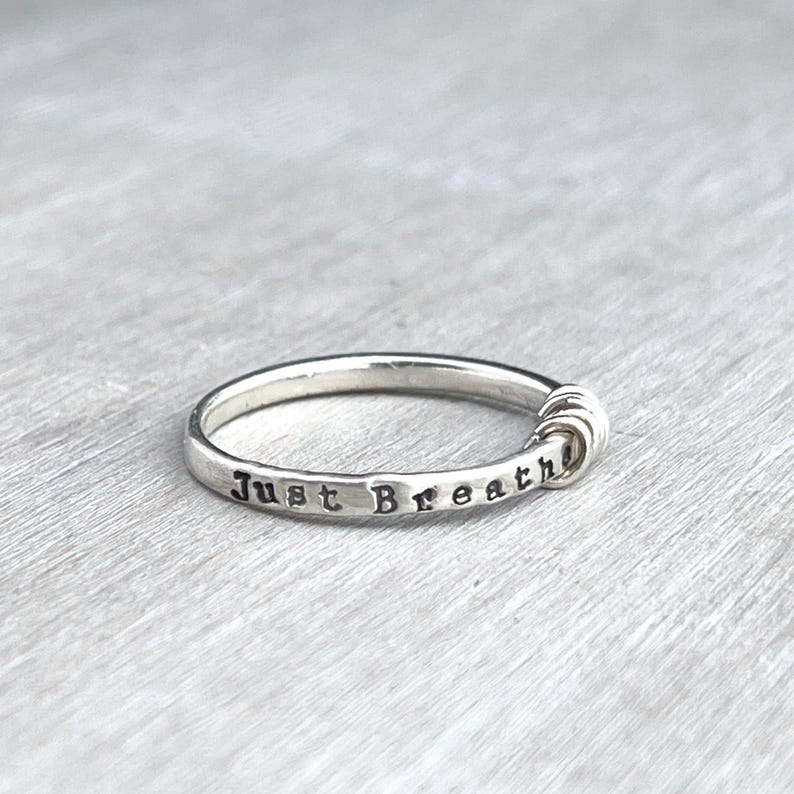 Pu&ograve; includere: Un anello d'argento con le parole "Just Breathe" incise. L'anello presenta un design minimalista con un elemento decorativo su un lato. L'anello &egrave; mostrato su una superficie grigio chiaro.